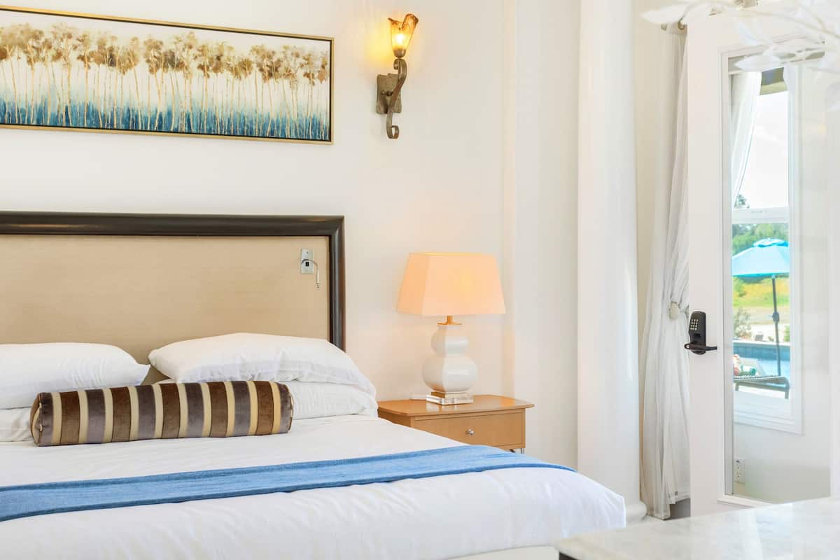 A hotel room with a bed, a nightstand with a lamp, a painting on the wall, and a door leading to an outdoor area.