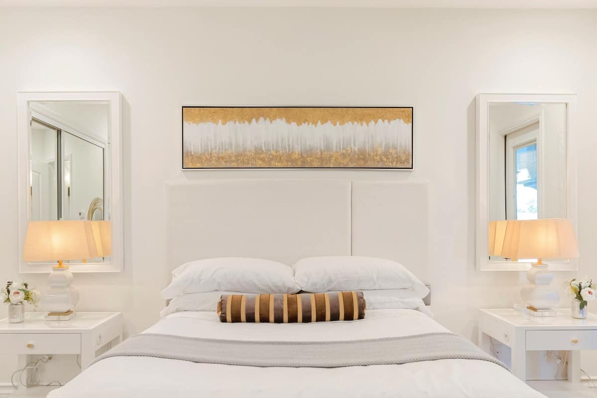 A modern bedroom with a large bed, two bedside tables with lamps, a mirror, and a wall-mounted painting.