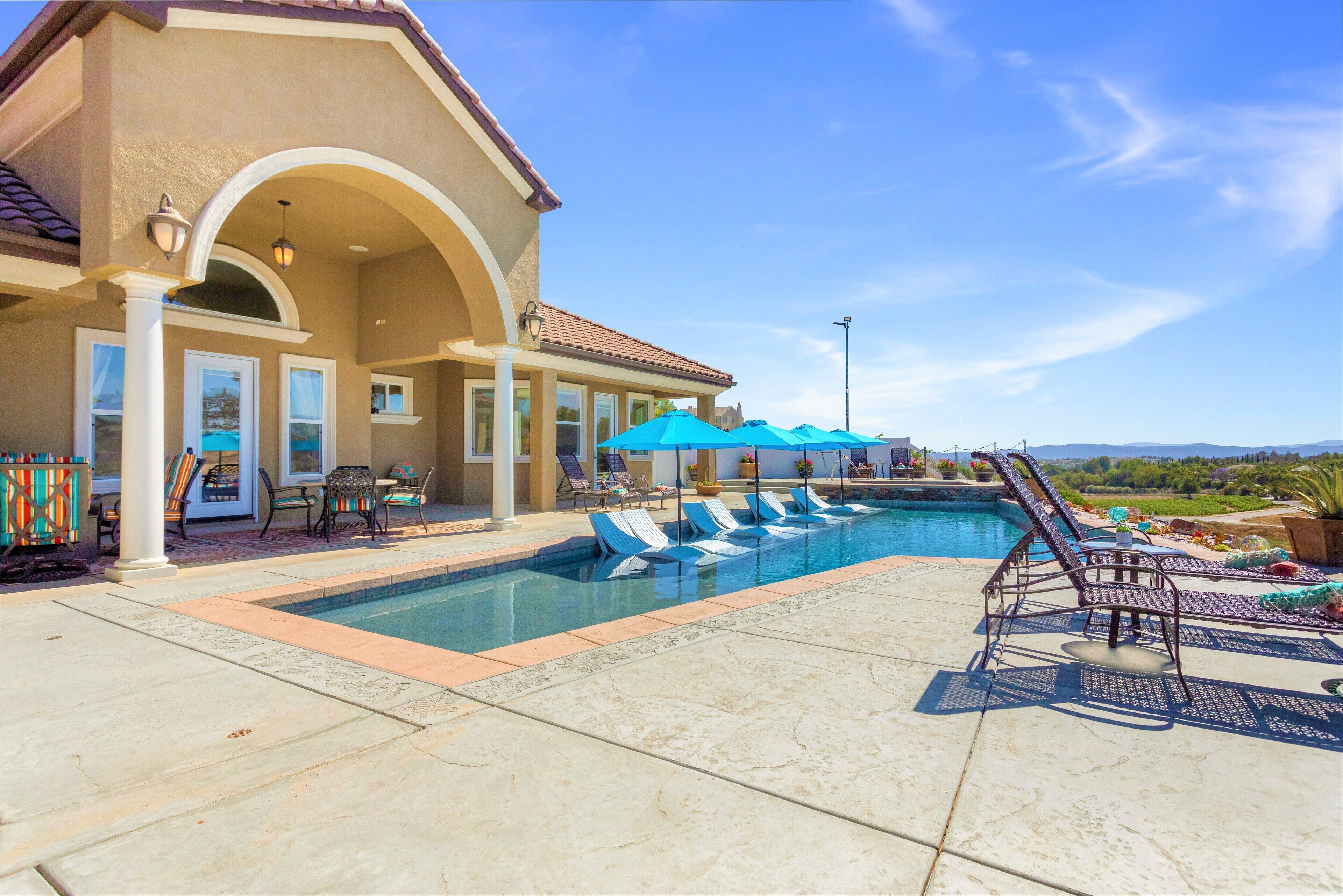A swimming pool with lounge chairs and umbrellas, a covered patio area, and a view of the landscape.