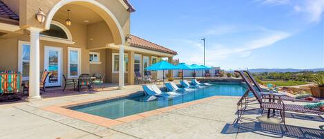 A swimming pool with lounge chairs and umbrellas, a covered patio area, and a view of the landscape.