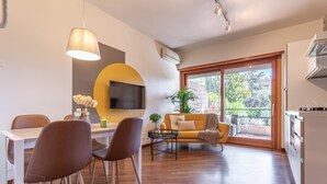 A modern living room with a sofa, dining table, and chairs. There is a TV mounted on the wall and a window with a view of greenery. - Oscar Garden Appartment   close to S. Pietro (ROMA)