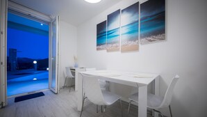 A dining area with a white table and chairs, a view through a glass door, and three paintings on the wall.
