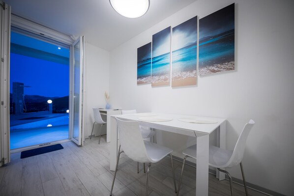 Un comedor con una mesa y sillas blancas, vistas a través de un ventanal y tres cuadros en la pared.