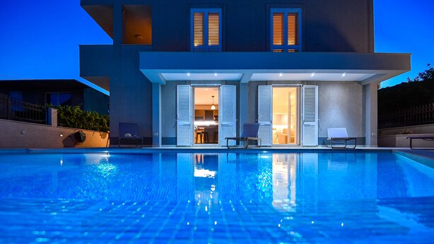 A modern house with a swimming pool, open doors leading to a dining area, and a well-lit exterior.