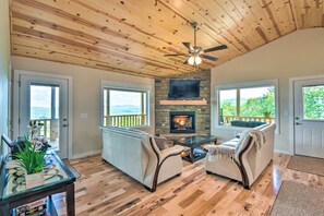 Relax in this spacious living area with a high ceiling, featuring a stone fireplace and large windows offering views of the outdoors.