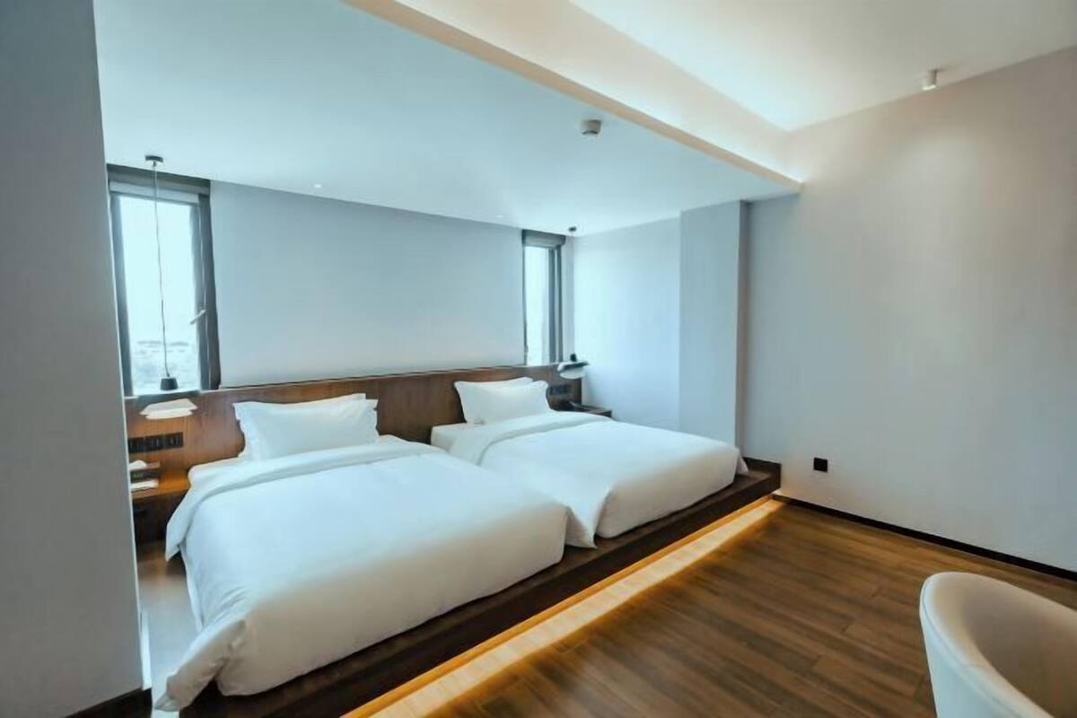 A modern hotel room with two beds, wooden flooring, and a large window providing natural light.
