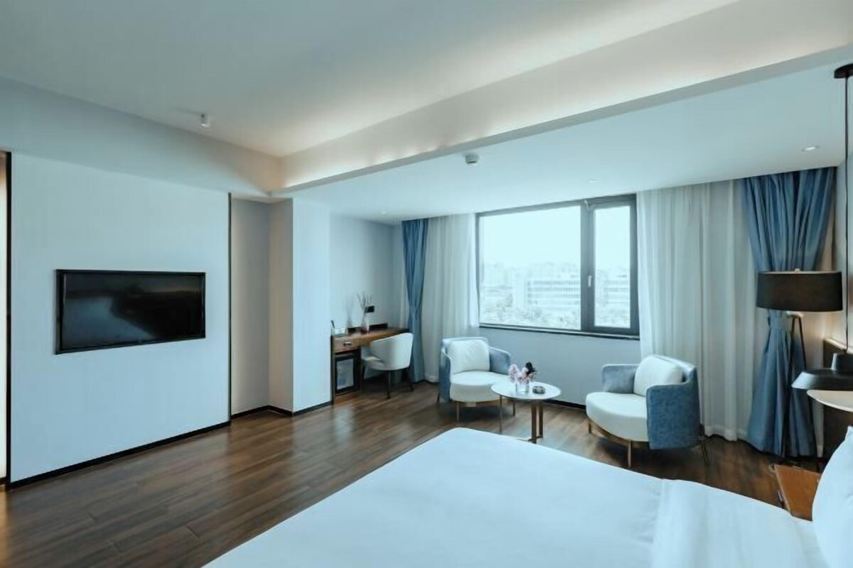 A modern hotel room with a large bed, a desk, two chairs, a TV, and a large window with curtains.