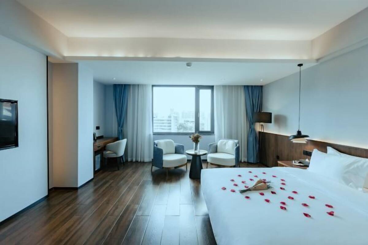 A hotel room with a large bed, two armchairs, a desk, and a view of the city through a large window.