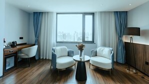 Una habitación de hotel moderna con suelo de madera, un escritorio con silla, dos sillones, una mesa de centro redonda y un gran ventanal con cortinas.