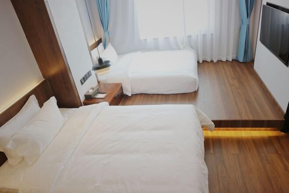 A hotel room with two beds, wooden flooring, a TV on the wall, and a window with curtains.