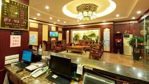 A hotel reception area with a digital clock, a reception desk with computers, a television, and a seating area with wooden furniture. - Feilong Holiday Hotel (Xinxing)