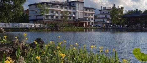A building with multiple floors, a body of water in the foreground, and greenery around.