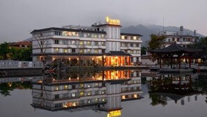 A multi-story hotel with a reflection in the water, a traditional pavilion, and mountains in the background. - Feilong Holiday Hotel (Xinxing)