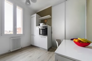 A modern kitchen with white cabinets, a built-in oven, and a microwave. There's a red bowl of fruit on the counter and a chair in the corner.