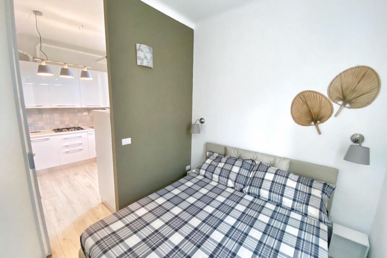 A bedroom with a bed, a nightstand, a wall-mounted lamp, and a view of a kitchen.