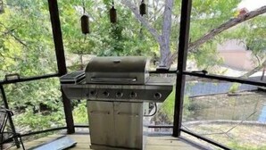 Ett takförsett utomhusområde med grillplats, trägolv och utsikt över träd och ett hus i bakgrunden.