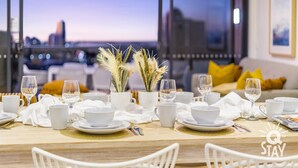 A dining table set for two with white tableware, a vase with dried plants, and a city view through large windows. - Ruby Apartments - Q Stay (Surfers Paradise)
