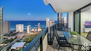 A balcony with a glass table, chairs, and a potted plant, overlooking a cityscape and the ocean. - Ruby Apartments - Q Stay (Surfers Paradise)