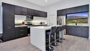 A modern kitchen with dark cabinets, a central island with bar stools, stainless steel appliances, and a large window with a view of a golf course. - Multi month discount,luxury,entertaining +relaxing (Cathedral City)