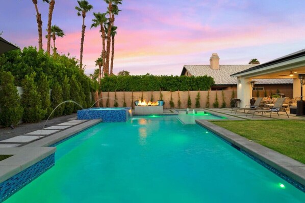 A swimming pool with a fire feature, surrounded by palm trees and a patio area.