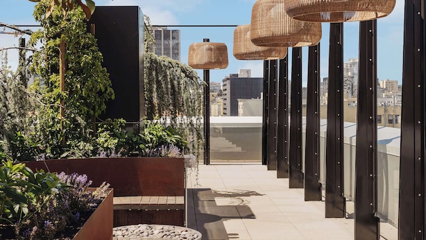 A rooftop terrace with potted plants, a wooden bench, and wicker pendant lights.