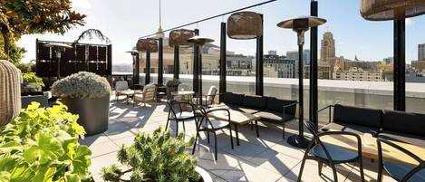 Eine Dachterrasse mit Sitzgelegenheiten im Freien, Topfpflanzen und Blick auf die Stadt.