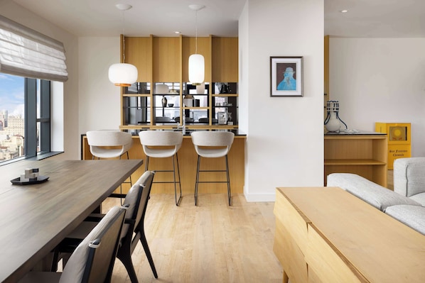 A modern kitchen with wooden cabinets, a dining area with a wooden table and chairs, and a view of the city through large windows. - Timbri Hotel San Francisco, Curio Collection by Hilton (San Francisco)