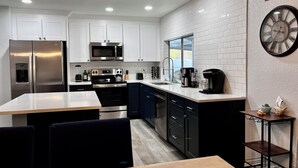 A modern kitchen with white cabinets, stainless steel appliances, a black countertop, and a dining area with a wooden table and blue chairs. - Free Heated Saltwater Pool, Kayaks, Bikes, Tennis Rackets and Fishing Poles! (North Port)