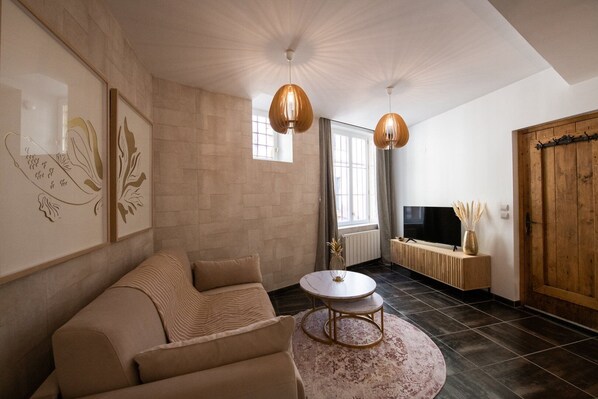 A living room with a sofa, a round coffee table, a TV on a wooden stand, and a large window with curtains.