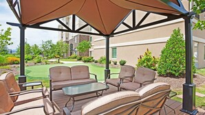 An outdoor patio with a table and chairs, a pergola with a brown canopy, and a view of a well-maintained lawn and buildings in the background. - Pool & Gym. Near University of Tennessee (Oak Ridge)