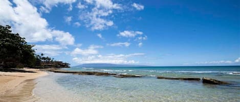 Une plage à l’eau claire, avec un rivage sablonneux et des affleurements rocheux.