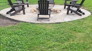 A circular outdoor seating area with four Adirondack chairs around a central fire pit, surrounded by a stone patio and a grassy lawn. - 2 Bedroom Coach House, Green Lake, WI (Green Lake)