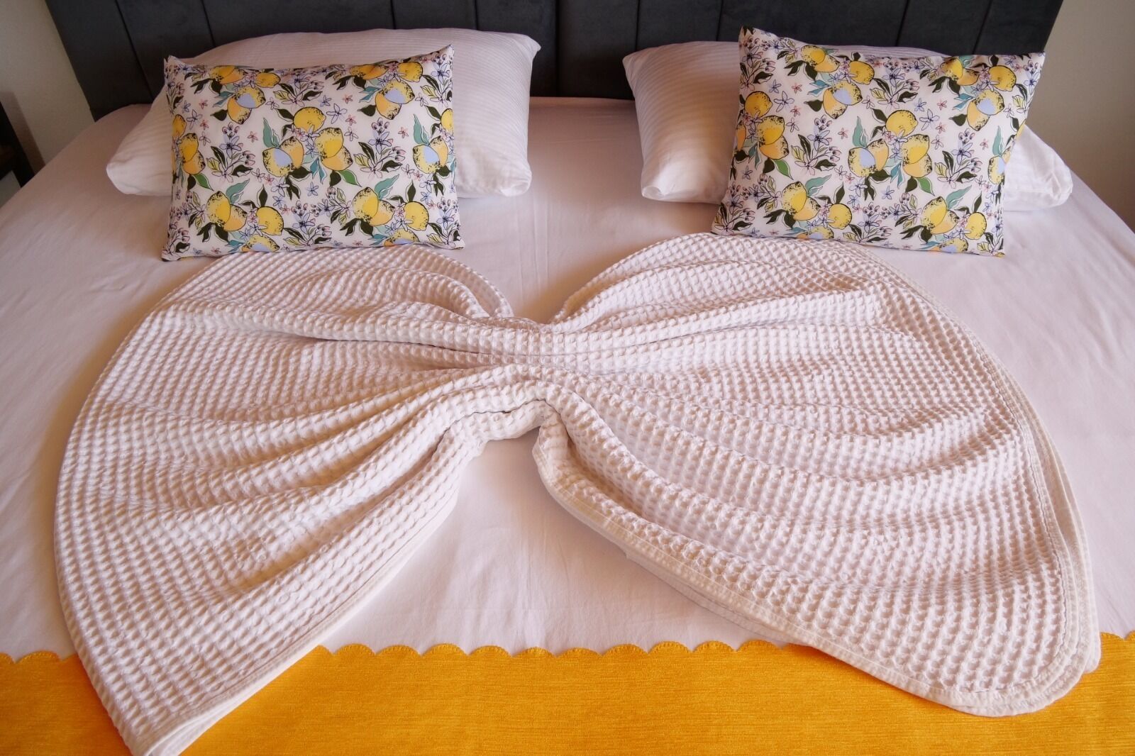 A bed with a large, textured white blanket and two pillows with a lemon and floral pattern.