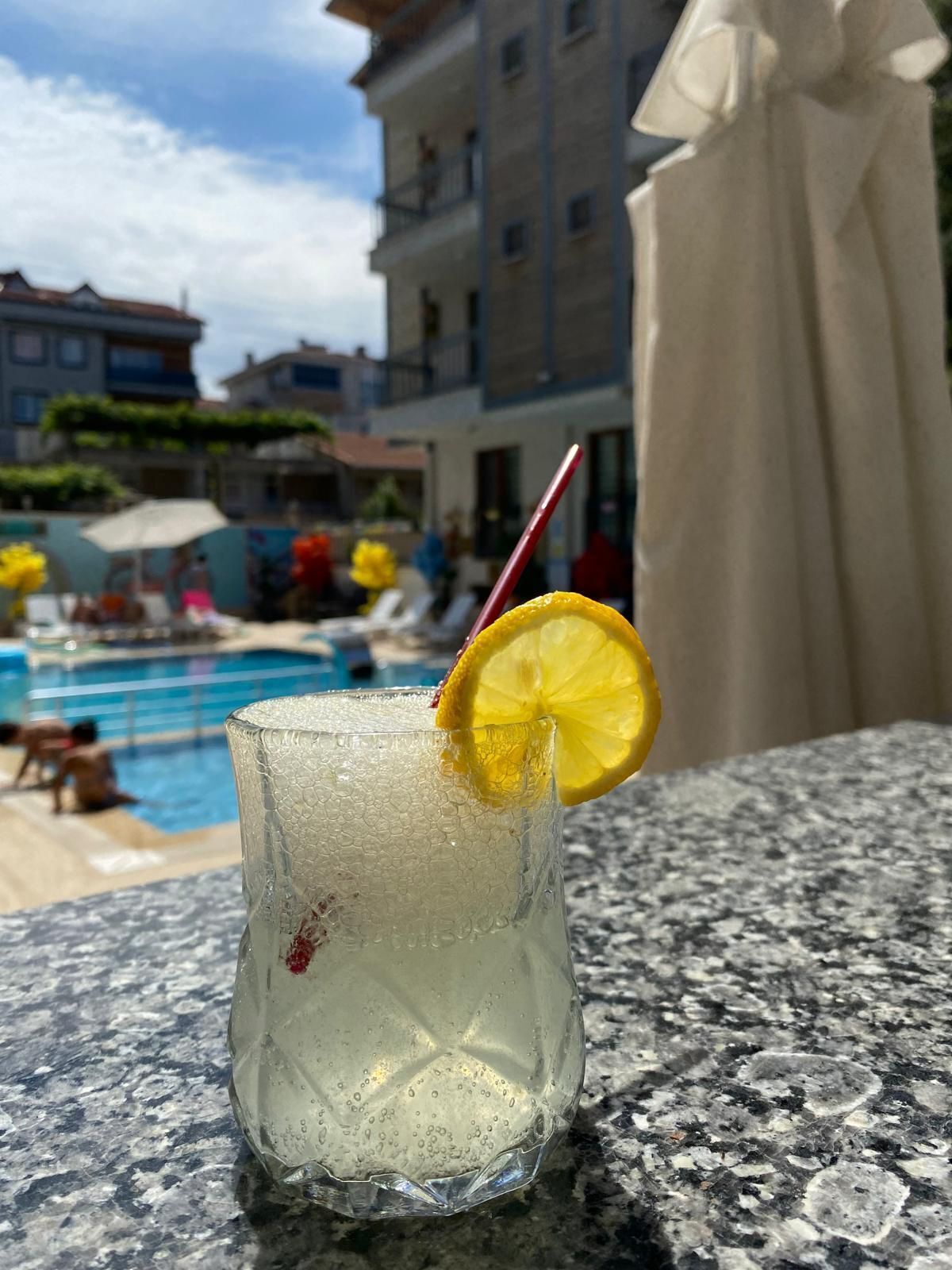 A glass of a clear beverage with a lemon slice garnish, placed on a granite surface near a poolside area with lounge chairs and umbrellas.