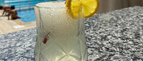 A glass of a clear beverage with a lemon slice garnish, placed on a granite surface near a poolside area with lounge chairs and umbrellas.