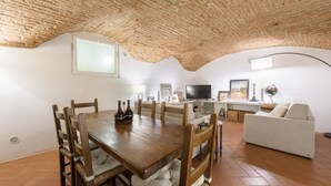 Zona pranzo con tavolo e sedie in legno, divano bianco e televisione in una stanza con soffitto a mattoni a vista e pavimento piastrellato.