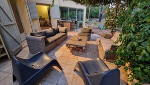 Ein möblierter Patio-Bereich im Freien mit Korbmöbeln, einem Holztisch und einer Glastür, die in einen Garten führt.