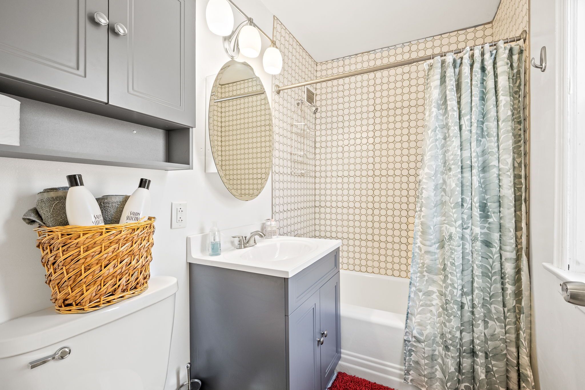 A bathroom with a white sink, grey cabinets, a wicker basket with toiletries, a mirror, and a shower with patterned curtains.