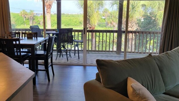 Une pièce dotée d’une porte-fenêtre coulissante qui donne sur un balcon offrant une vue sur la verdure et un paysage lointain. À l’intérieur, on trouve un canapé et une table à manger avec des chaises.
