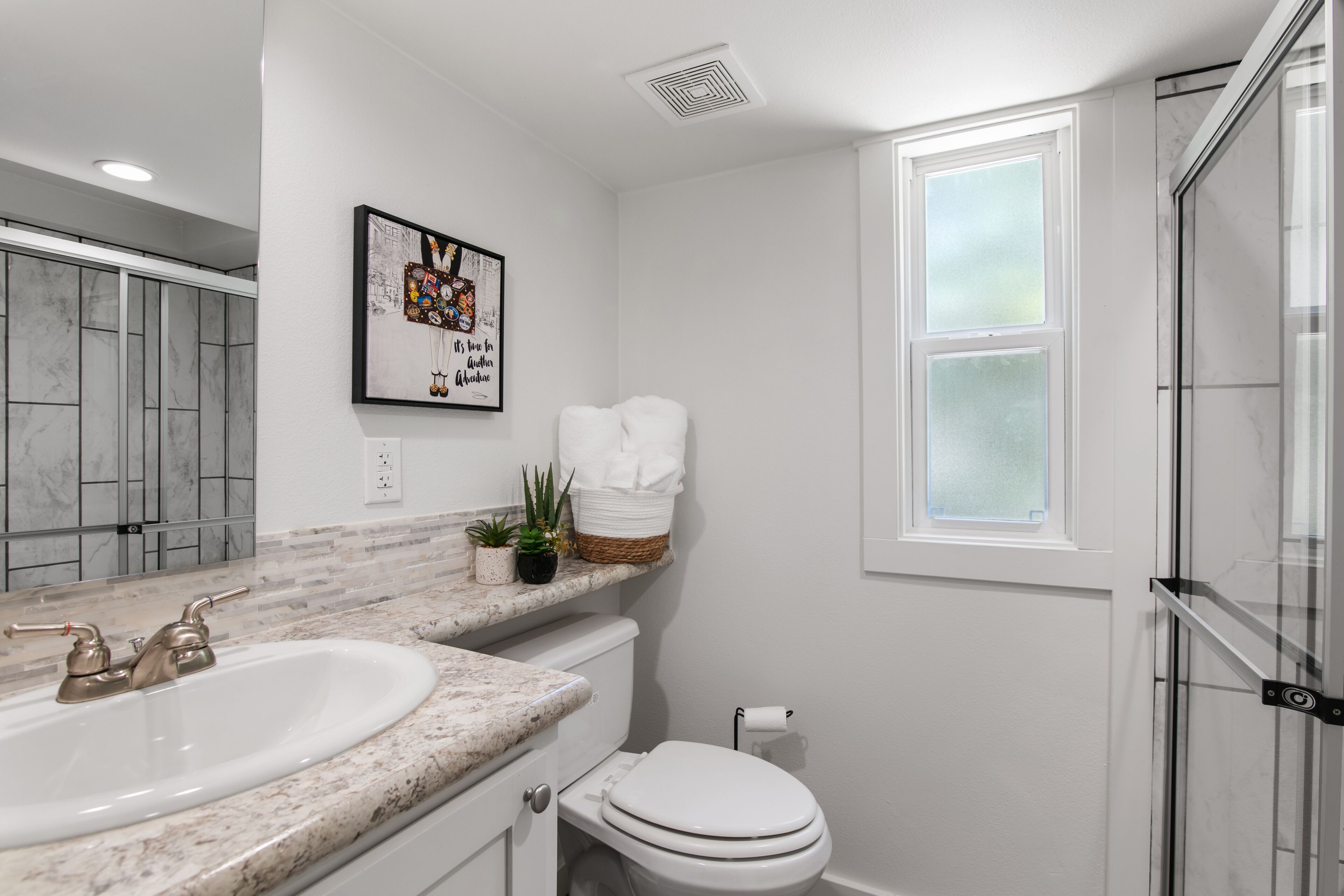 A bathroom with a double sink, a toilet, a shower, and a window.