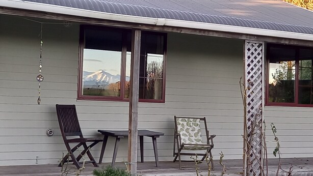 Un bâtiment doté d’un toit en métal, de deux fenêtres donnant sur une montagne et d’un porche avec une table et deux chaises.