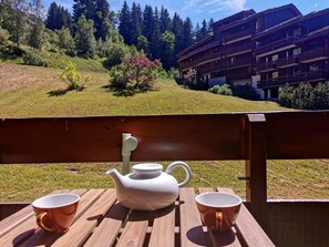 A wooden table with a white teapot and two mugs, set against a backdrop of a mountainous landscape and a chalet-style building.