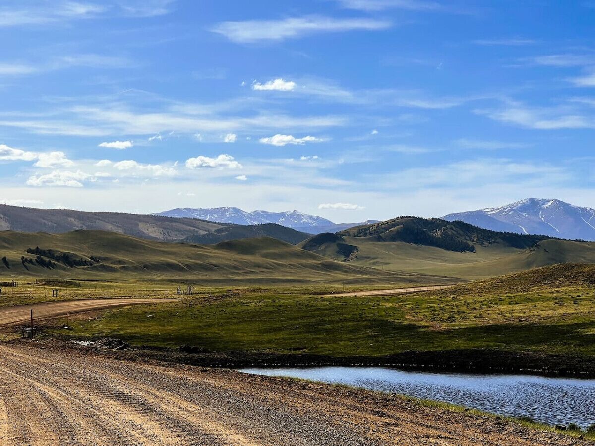 A rural landscape with a dirt road, a small water body, and distant snow-capped mountains under a clear sky.