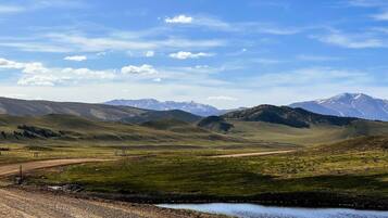A rural landscape with a dirt road, a small water body, and distant snow-capped mountains under a clear sky.