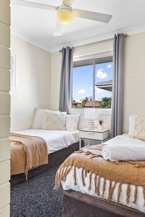 Een hotelkamer met twee bedden, een plafondventilator, een raam met gordijnen en uitzicht op het buiten.