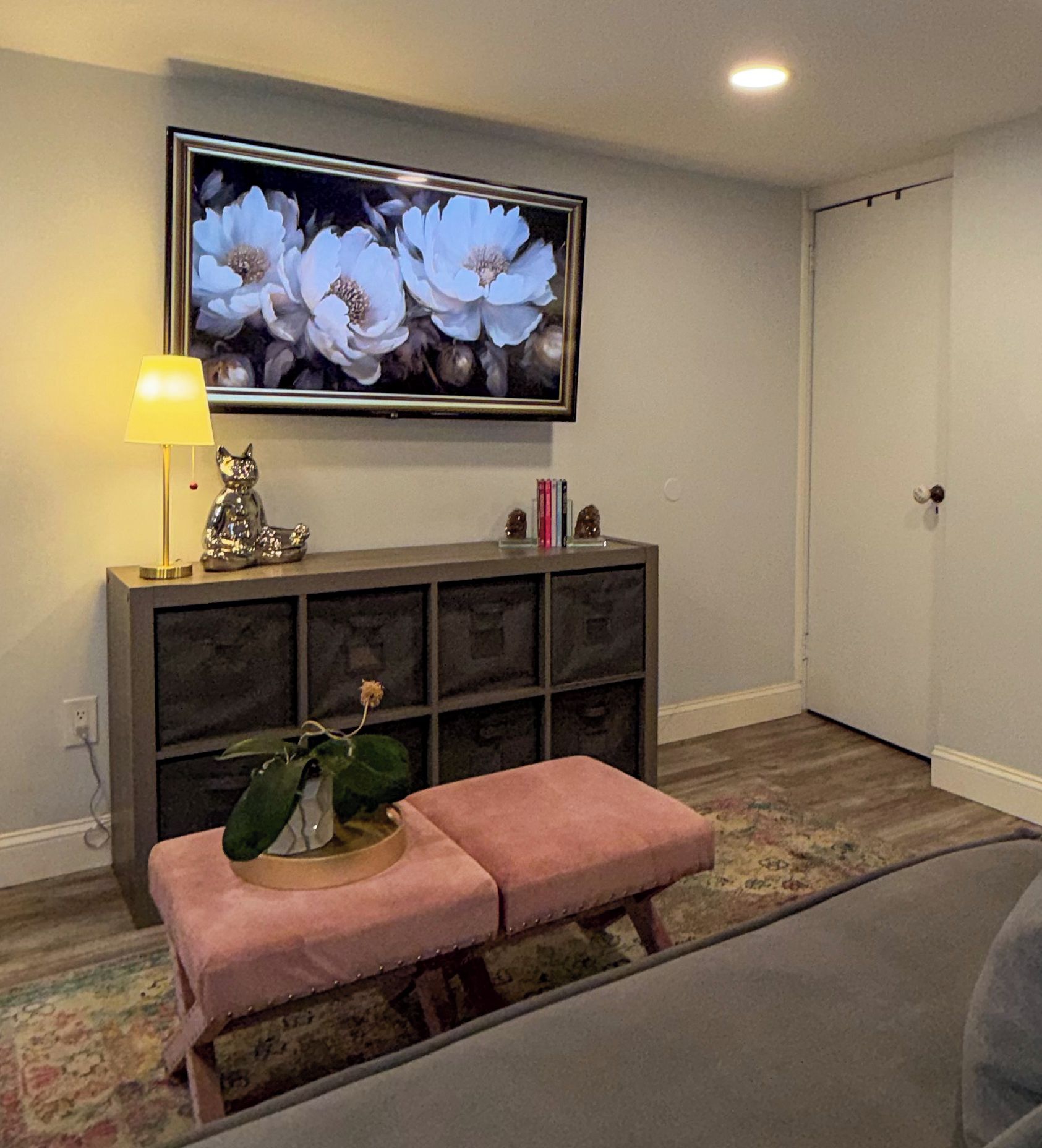 A living room with a television mounted on the wall, a bench, a lamp, and a bookshelf.