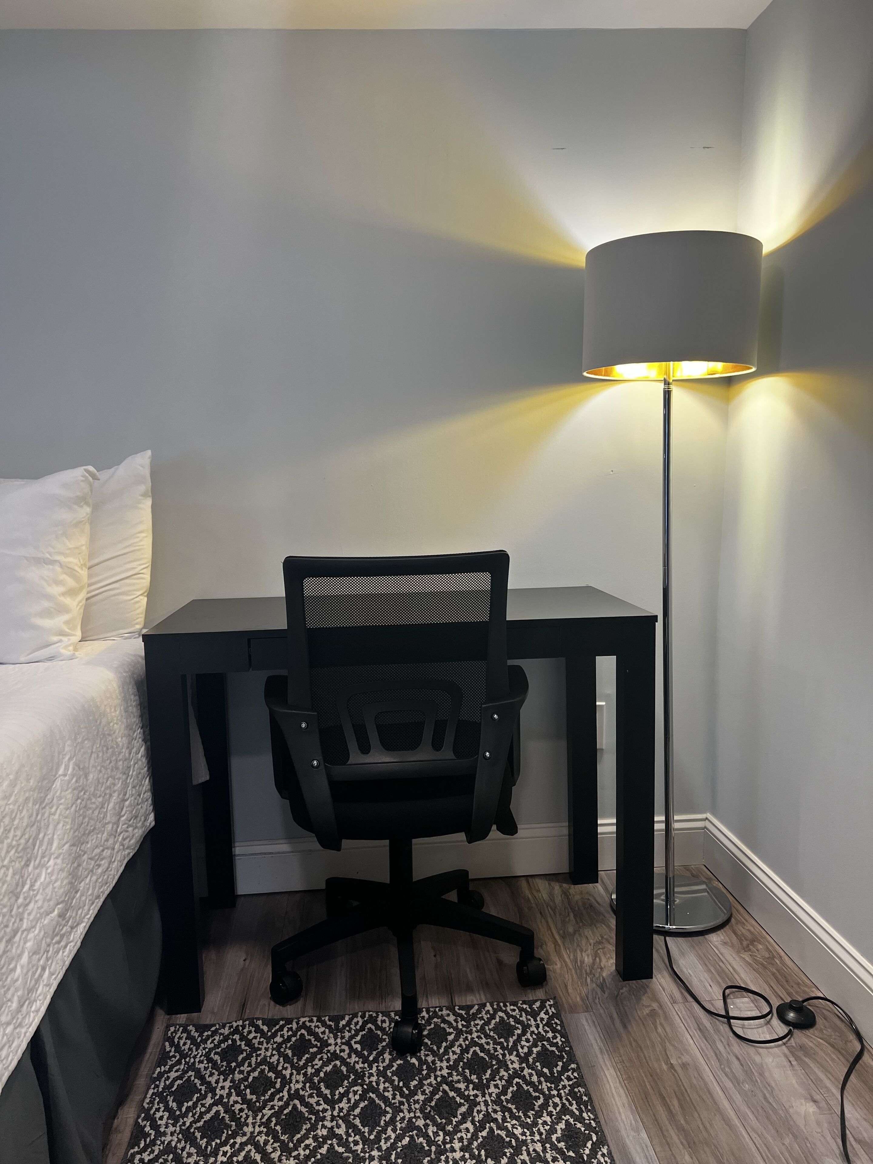 Een slaapkamer met een bed, een bureau, een stoel, een vloerlamp en een kamerkleed met een patroon.
