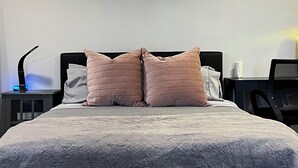 Un lit avec une couette grise et deux oreillers roses, une tête de lit noire et une petite table de chevet avec une lampe.