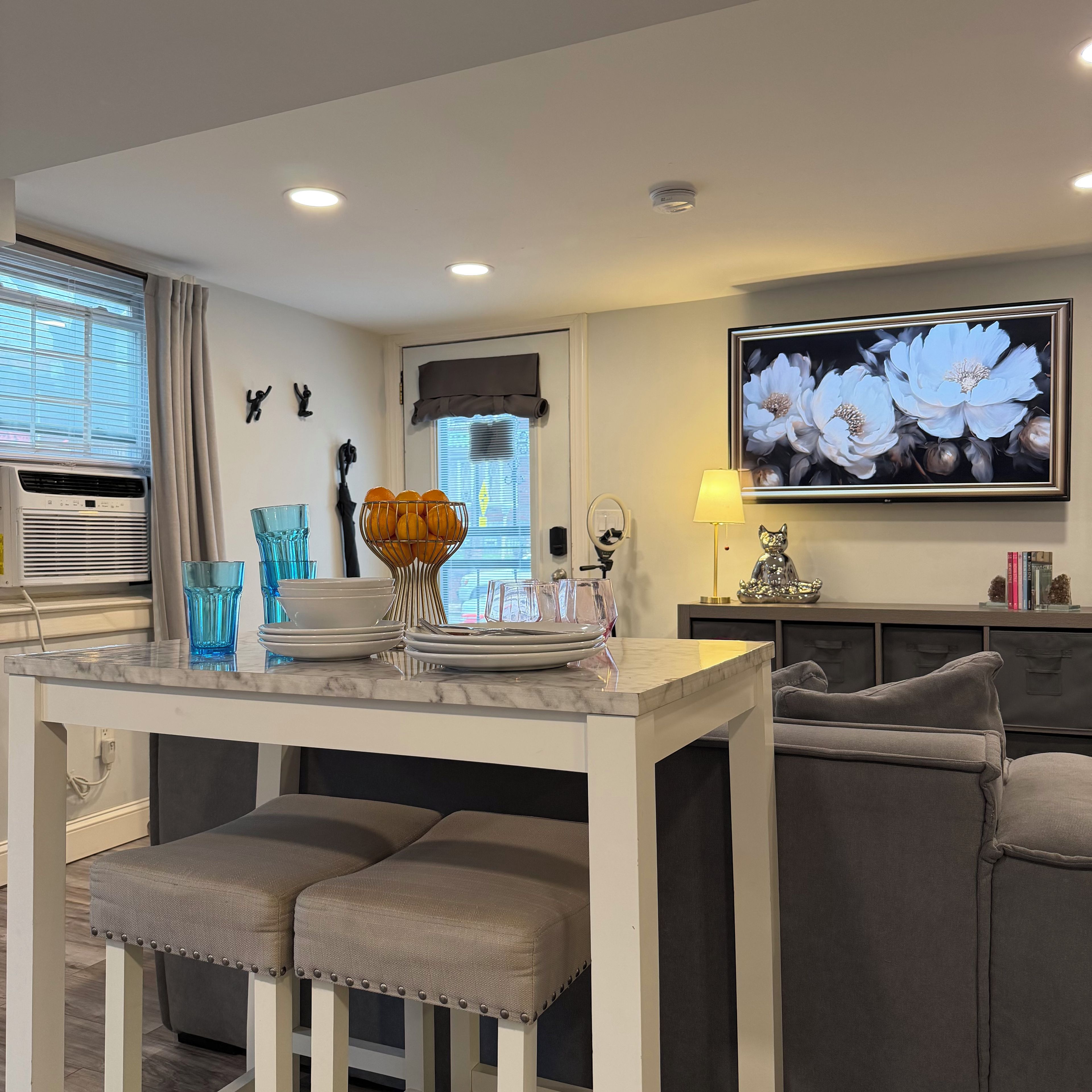 A modern kitchen with a marble countertop, a dining area with two chairs, a wall-mounted TV, and a window with blinds.