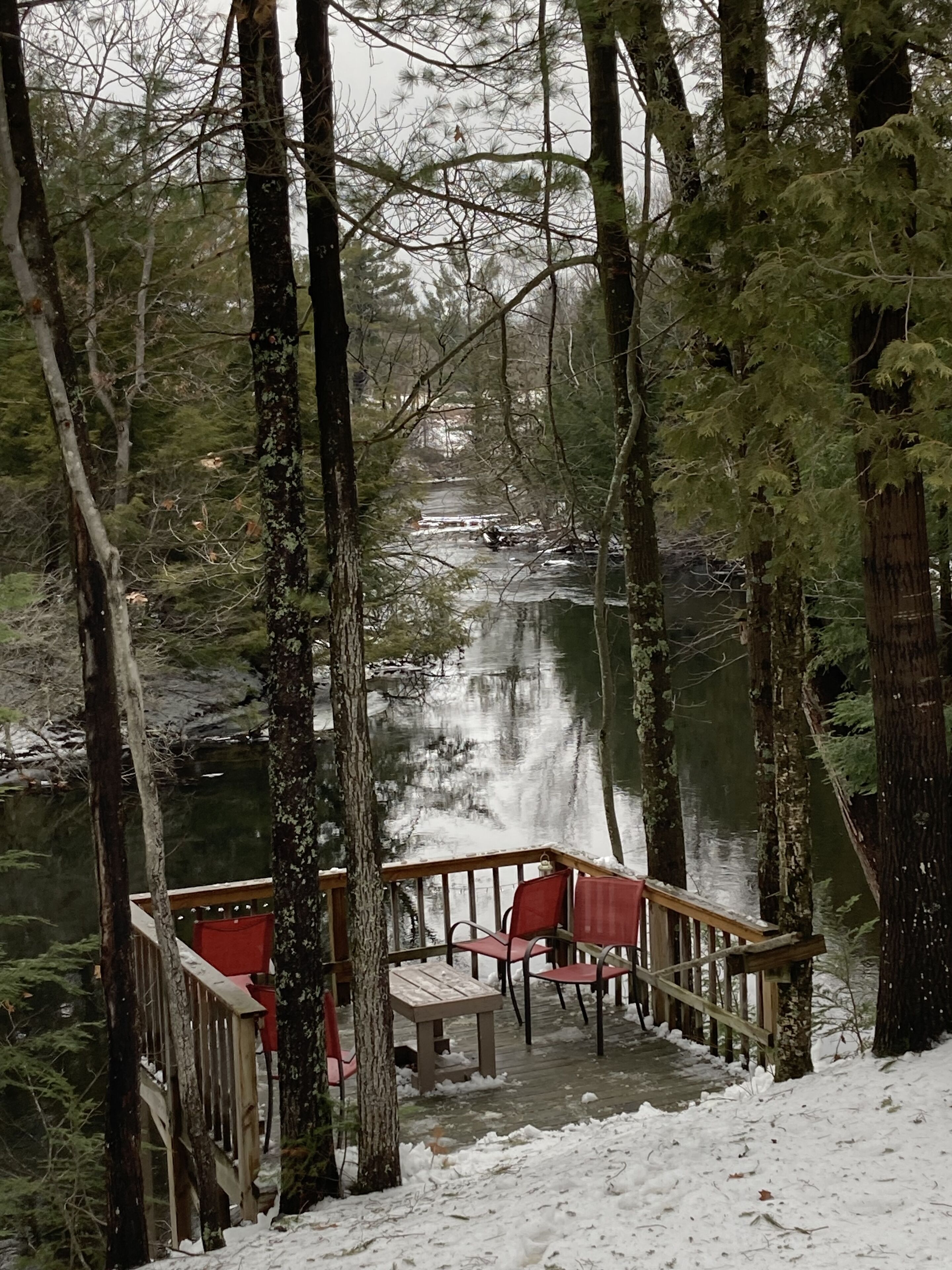 A wooden deck with chairs and a table overlooking a river, surrounded by tall trees.